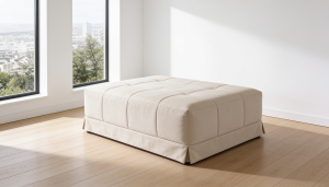 Puff cama para invitados tapizado en color beige, con diseño de cuadrados acolchados y faldón en la base, ubicado en un salón moderno con suelo de madera y ventanas grandes.